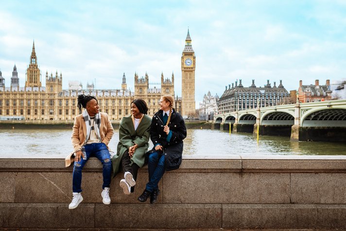 A Black man with dreadlocks, a Black woman, and a white woman with red hair, laughing and smiling while sitting on a wall next to the River Thames, with Parliament on the opposite bank of the river.